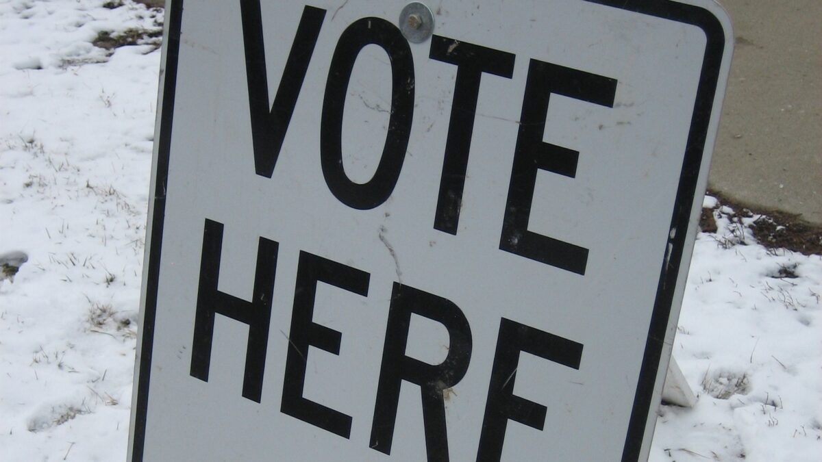 A white Vote Here sign on a stand in snowy grass beside a sidewalk outside a Michigan polling place.