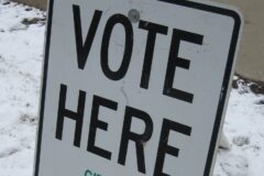 A white Vote Here sign on a stand in snowy grass beside a sidewalk outside a Michigan polling place.