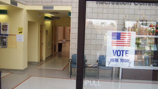 A Vote Here Today sign with an American flag taped to the glass door of a New Jersey polling place in a recreation center lobby.