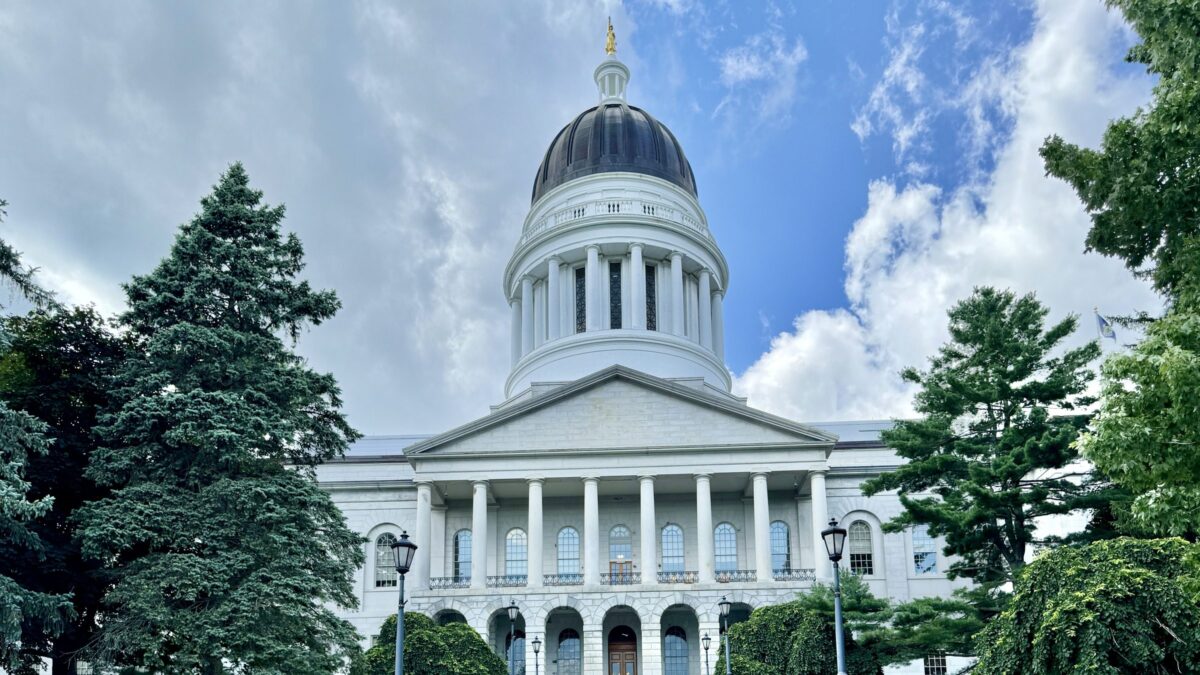 The Maine State House in Augusta, with its dome and columned granite portico framed by evergreen trees under a partly cloudy sky.