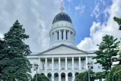 The Maine State House in Augusta, with its dome and columned granite portico framed by evergreen trees under a partly cloudy sky.