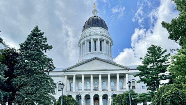 The Maine State House in Augusta, with its dome and columned granite portico framed by evergreen trees under a partly cloudy sky.